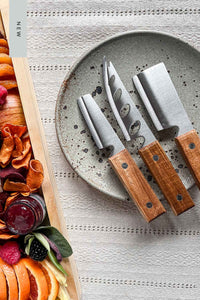 A set of 3 rustic acacia wood cheese knives on a speckled pottery plate next to a cheese board.