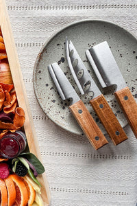 A set of 3 rustic acacia wood cheese knives on a speckled pottery plate next to a cheese board.