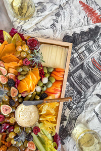 The Après Ski Cheese Board filled with Vermont and alpine cheese, pickles, fruits, vegetables, and nuts on a table surrounded by wine glasses.