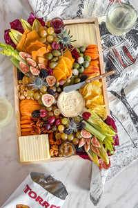 The Après Ski Cheese Board filled with Vermont and alpine cheese, pickles, fruits, vegetables, and nuts on a table surrounded by wine glasses and truffle potato chips.