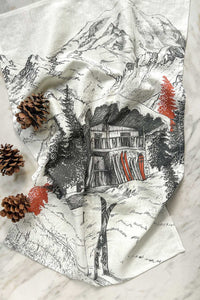 A printed black, white, and red linen tea towel with an après ski scene of a snowy mountain cabin chalet surrounded by evergreen trees and skis. 