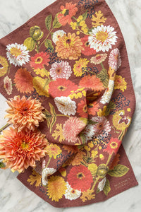 An autumnal hued tea towel with fall flowers, leaves, and berries with peach dahila flowers on a marble counter.