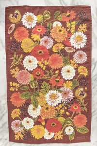 An autumnal hued tea towel with fall flowers, leaves, and berries on a marble counter.