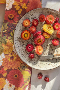 An autumnal hued tea towel with fall flowers, leaves, and berries with strawflowers and a speckled plate on a marble counter.