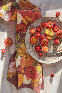 An autumnal hued tea towel with fall flowers, leaves, and berries with strawflowers and a speckled plate on a marble counter.