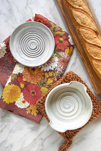 A handmade ivory stoneware brie baker and garlic grater sitting on a marble countertop with a wool trivet and an autumnal linen tea towel.