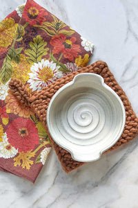 A handmade ivory stoneware brie baker sitting on a wool trivet next to a linen tea towel in an autumnal floral design.