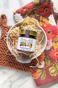 A handmade ivory stoneware brie baker sitting on a wool trivet next to a linen tea towel in an autumnal floral design. Paired with Caramelized Fig Preserves with Pear & Honey