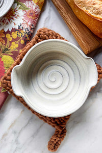 A handmade ivory stoneware brie baker sitting on a wool trivet.