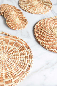 An assorted display of handmade longleaf pine needle trivets and coasters in three different patterns on a white marble counter.