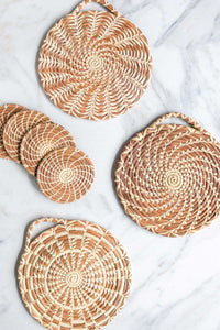 A set of four handmade longleaf pine needle coasters and three assorted design trivets on a white marble counter.