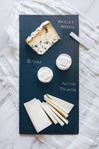 A slate cheese board with an assortment of cheeses, the cheese names are written on the slate with chalk.