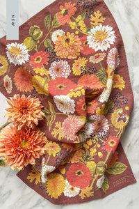 A fall toned tea towel in warm oranges, peaches, gold, and brown with a design of fall flowers, berries, and leaves, laid next to peach dahlia flowers.