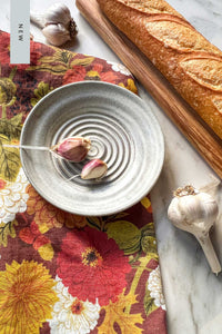 A handmade stoneware garlic grater dipping plate with two garlic cloves in it sits on an autumnal floral tea towel next to a crusty baguette on a maple wood serving board.