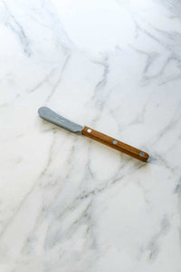 The Bistrot Spreader Knife, In Vintage Teak lying on a marble counter.