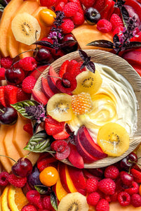 A ready-to-eat seasonal fruit platter featuring fresh melons, stone fruit, berries, kiwi, and edible flowers on a marble counter, available for Vermont delivery.