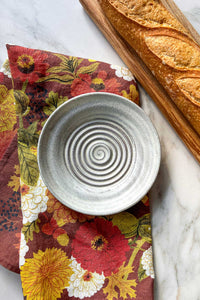 A handmade stoneware garlic grater dipping plate sits on an autumnal floral tea towel next to a crusty baguette on a maple wood serving board.