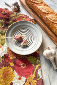 A handmade stoneware garlic grater dipping plate with two garlic cloves in it sits on an autumnal floral tea towel next to a crusty baguette on a maple wood serving board.