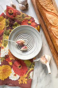 A handmade stoneware garlic grater dipping plate with two garlic cloves in it sits on an autumnal floral tea towel next to a crusty baguette on a maple wood serving board.