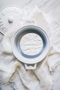 A ceramic brie baker with a brie cheese inside waiting to be baked. Sitting on top of an ivory linen napkin.