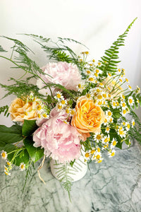 The Floral Garden Bouquet is a mix of soft pastel garden flowers, ferns, and greens arranged in a white ceramic vase.
