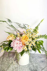 The Floral Garden Bouquet is a mix of soft pastel garden flowers, ferns, and greens arranged in a white ceramic vase.