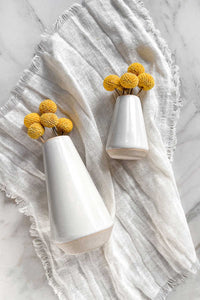 A pair of white stoneware bud vases with raw clay edges and bases, lying on an ivory linen napkin.
