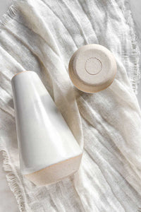 A medium white stoneware bud vase with a raw clay edge and base, lying on an ivory linen napkin.