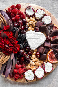 A large round cheese board filled with chocolate, cheese, fresh fruits, nuts, and flowers.