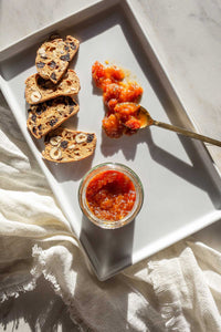 A white porcelain platter with an open jar of Peach Bulgarian Pepper Tomato Honey Jam with a spoonful smeared on the tray next to crackers.