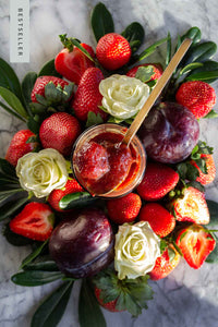 Piles of fresh strawberries, white roses, and ripe plums surround a jars of Strawberry Italian Plum Rosewater Honey Jam.