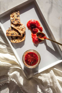 A white porcelain platter with an open jar of Strawberry Italian Plum Rosewater Honey Jam with a spoonful smeared on the tray next to crackers.